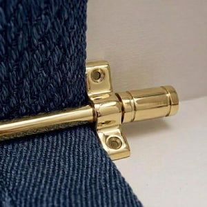 May include: Close-up of a polished brass door bolt. The bolt is mounted on a textured navy blue fabric. The bolt has a cylindrical body and a rectangular mounting plate with screw holes. The background is a light beige.