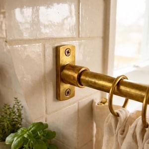 May include: A brass curtain rail with rings, fixed to a cream-coloured tiled wall. The rail is attached with a rectangular brass bracket. The scene includes a glimpse of a window and some potted herbs.