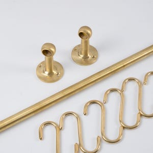 Puede incluir: Un conjunto de componentes de ferretería de latón, que incluye una barra larga, dos soportes de montaje y varios ganchos en forma de S. Los artículos están dispuestos sobre una superficie blanca, mostrando su acabado metálico.