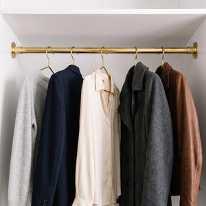 May include: A brass closet rod mounted between white walls, holding five hangers with a grey sweater, a navy blazer, a cream button-down shirt, a charcoal grey wool coat, and a brown leather jacket, all displayed in a clean, organized fashion.