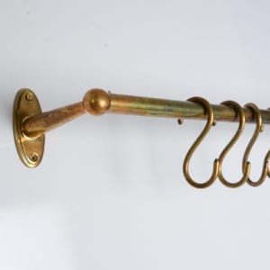 May include: A wall-mounted brass rail featuring a horizontal rod with rounded ends and two oval-shaped mounting plates. Four matching brass S-shaped hooks are suspended from the rod, providing a functional storage solution for hanging various items.