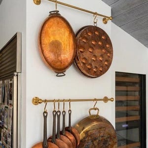 Peut inclure: Batterie de cuisine en cuivre suspendue à des barres en laiton contre un mur blanc. L'ensemble comprend des casseroles ovales, une poêle ronde à rivets et plusieurs poêles en cuivre avec de longues poignées. Décor de cuisine.