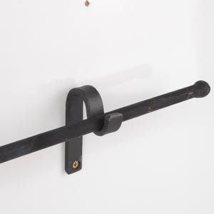 May include: A black, wrought iron towel bar mounted on a white wall. The bar has a rounded end and is held in place by a curved bracket. The fixture is designed for a bathroom or kitchen.