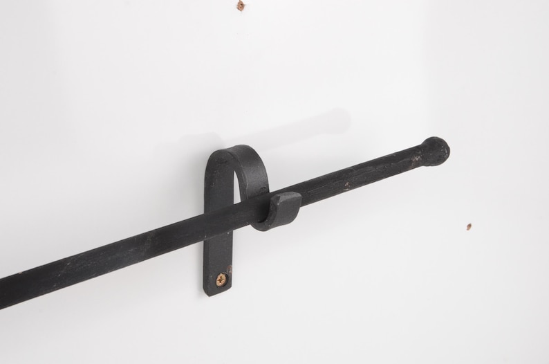 May include: A black, wrought iron towel bar mounted on a white wall. The bar has a rounded end and is held in place by a curved bracket. The fixture is designed for a bathroom or kitchen.