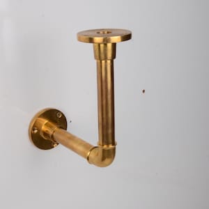 May include: A brass wall-mounted shelf bracket. The bracket has a circular base, a straight vertical pipe, and a horizontal pipe connected by a 90-degree elbow. The top has a circular plate. The hardware is gold-colored.