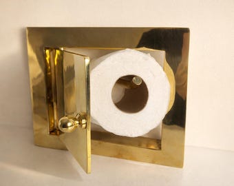 May include: A gold-colored, rectangular toilet paper holder with a hinged door and a round knob. The holder is mounted on a wall and contains a roll of white toilet paper.