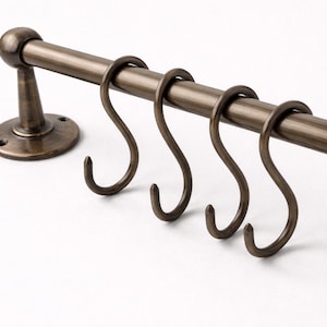 May include: Bronze-toned metal kitchen rail with four S-shaped hooks. The rail is mounted on a round base with screw holes and a decorative support. The hooks are evenly spaced along the rail, offering a classic storage solution.