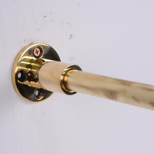 May include: A brass curtain rod with a circular mounting plate attached to a white wall. The rod is cylindrical and extends horizontally from the mounting plate. The mounting plate has screw holes and is secured with screws.