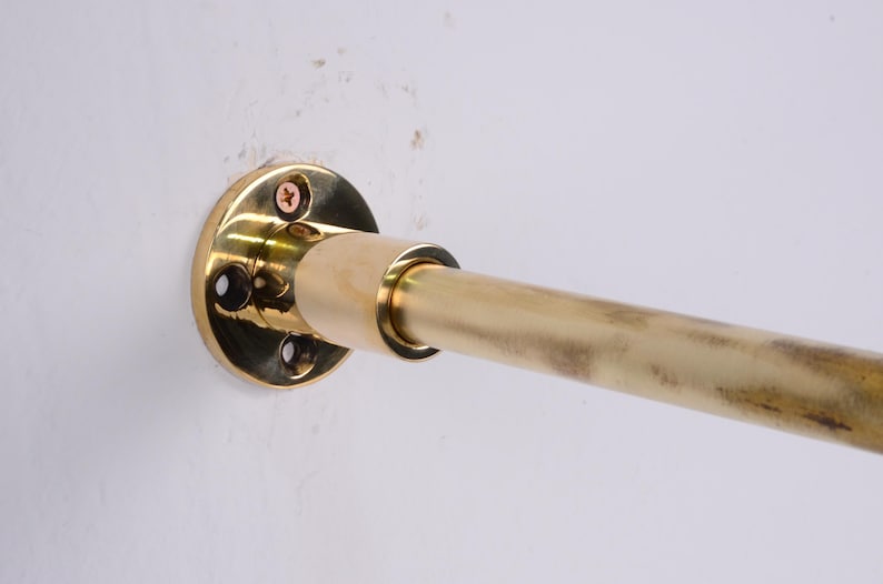 May include: A brass curtain rod with a circular mounting plate attached to a white wall. The rod is cylindrical and extends horizontally from the mounting plate. The mounting plate has screw holes and is secured with screws.