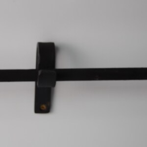 May include: A black, wrought iron towel bar with a rounded end. The bar is mounted on a vertical bracket with a square center and a small, round detail. The design is simple and functional.