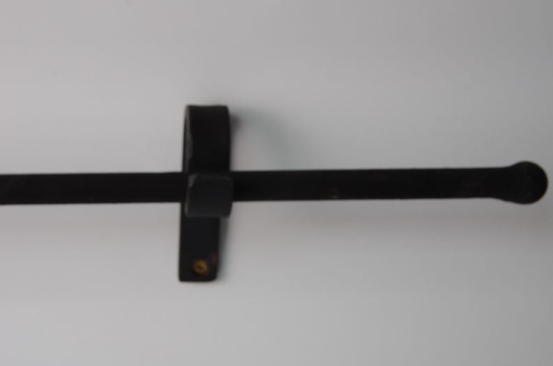 May include: A black, wrought iron towel bar with a rounded end. The bar is mounted on a vertical bracket with a square center and a small, round detail. The design is simple and functional.