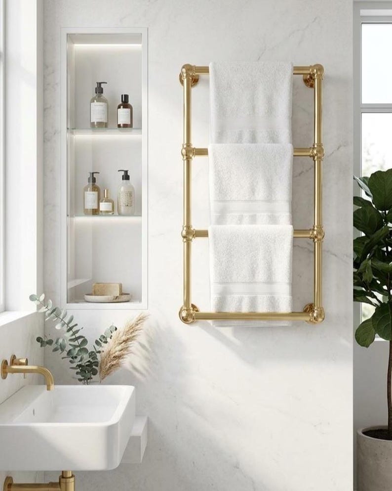 May include: A gold-colored towel rack with three white towels hanging on it. The rack is mounted on a white marble wall in a bathroom setting. The bathroom also features a white sink, a built-in shelf with toiletries, and a potted plant.