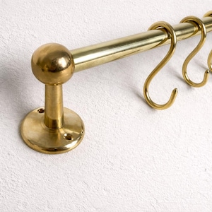 May include: A brass kitchen rail with four S-hooks. The rail is mounted on a white wall. The rail has a round finial on one end and a circular base. The hooks are curved and designed for hanging items.