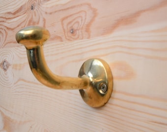 May include: A brass hook with a rounded top and a curved arm. The hook is mounted on a light wood surface.