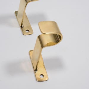 Könnte beinhalten: Zwei messingfarbene, gebogene Metallhaken. Jeder Haken hat eine rechteckige Basis mit einem einzelnen Loch zur Befestigung und ein geschwungenes, wellenartiges Design. Die Haken werden vor einem weißen Hintergrund gezeigt.