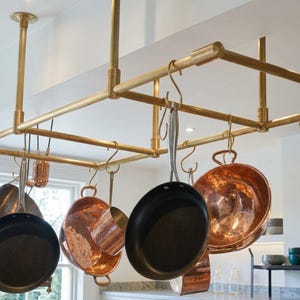 Op de afbeelding: Een aan het plafond gemonteerd pannenrek van messing met een rechthoekig frame en meerdere S-vormige haken. Verschillende koperen potten en pannen met een metallic afwerking en één zwarte koekenpan met antiaanbaklaag hangen aan de haken.