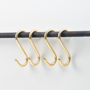 Pode incluir: Quatro ganchos em forma de S dourados pendurados numa barra de metal cinza escuro. Os ganchos são espaçados uniformemente e têm um design suave e curvo. O fundo é uma parede branca lisa.