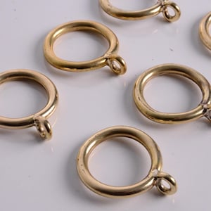 May include: Several gold-colored curtain rings with a loop for hanging. The rings are circular and have a smooth, metallic finish. The image shows the rings scattered on a white surface, suggesting a hardware or home decor theme.