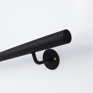 May include: A black metal handrail, fixed to a white wall. The cylindrical handrail has a curved support attached to a circular wall mount. The wall mount features a small, gold-coloured screw.