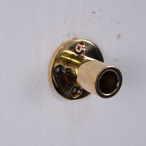 May include: A brass-colored wall-mounted pipe holder with a circular base and a cylindrical opening. The holder is attached to a white wall with screws.