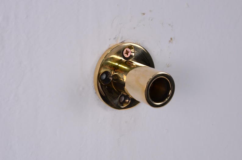 May include: A brass-colored wall-mounted pipe holder with a circular base and a cylindrical opening. The holder is attached to a white wall with screws.