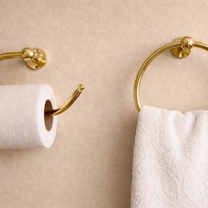 May include: A gold-colored toilet paper holder with a roll of white toilet paper and a gold towel ring with a white towel hanging on a beige wall. Bathroom decor.