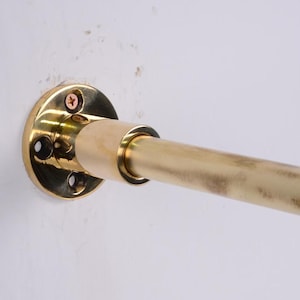 May include: A brass rod and mounting hardware are attached to a white wall. The rod is cylindrical and has a slightly aged appearance. The mounting hardware is circular with screw holes.