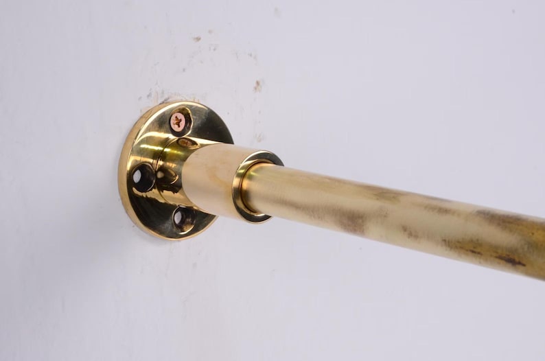 May include: A brass rod and mounting hardware are attached to a white wall. The rod is cylindrical and has a slightly aged appearance. The mounting hardware is circular with screw holes.