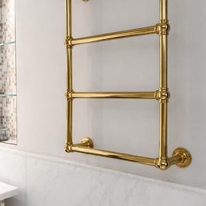 May include: A gold-colored, wall-mounted towel warmer with four horizontal bars. The design features rounded joints and circular wall mounts, creating a classic, elegant look. The warmer is installed on a white wall.