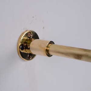 May include: A brass-colored curtain rod with a wall mount. The rod is cylindrical and has a slightly aged appearance. The wall mount is circular with screw holes for installation. The rod is mounted on a white wall.
