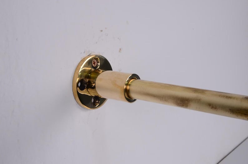 May include: A brass-colored curtain rod with a wall mount. The rod is cylindrical and has a slightly aged appearance. The wall mount is circular with screw holes for installation. The rod is mounted on a white wall.