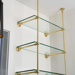 Op de afbeelding: Een wandgemonteerde stellingkast met drie niveaus, voorzien van heldere glazen planken ondersteund door een frame van gepolijst messing. De structuur bevat verticale en horizontale messing stangen die met ronde montageplaten aan de muur en het plafond zijn bevestigd.