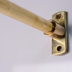 May include: A brass closet rod with a matching bracket, mounted on a white wall. The rod is cylindrical and appears to be made of wood. The bracket is rectangular with screw holes.