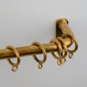 May include: A vintage brass curtain rail with matching rings. The rail is mounted on a white wall. The rail and rings have a warm, golden hue, suggesting an antique or retro style. The rings are designed to hold curtains.
