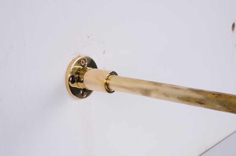 May include: A brass-colored, wall-mounted rod with a circular mounting plate. The rod is attached to the wall with screws. The rod is approximately 1 inch in diameter and extends horizontally from the wall.