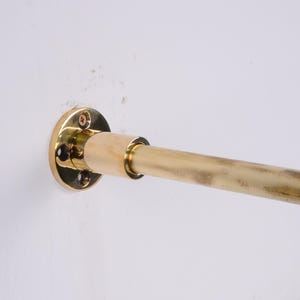 May include: A brass-coloured curtain rod is mounted on a white wall. The rod is cylindrical and extends horizontally from a circular mounting plate. The mounting plate is secured to the wall with screws.