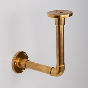 May include: A brass wall-mounted shelf bracket with a circular base and a right-angle design. The bracket features a vertical pipe connected to a horizontal pipe, with a circular top for holding a shelf. The brass has a polished finish.