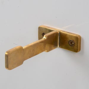 May include: A brass shelf bracket with a single screw visible. The bracket is designed to be mounted to a wall and has a flat surface for a shelf to rest on.