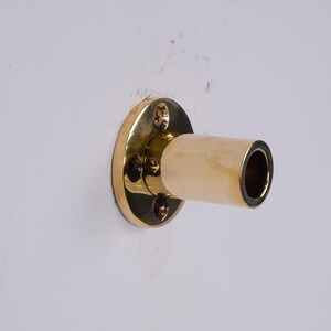 May include: A brass-colored wall-mounted curtain rod holder. The holder has a circular base with four screws and a cylindrical extension. The extension is a light beige color with a dark interior.