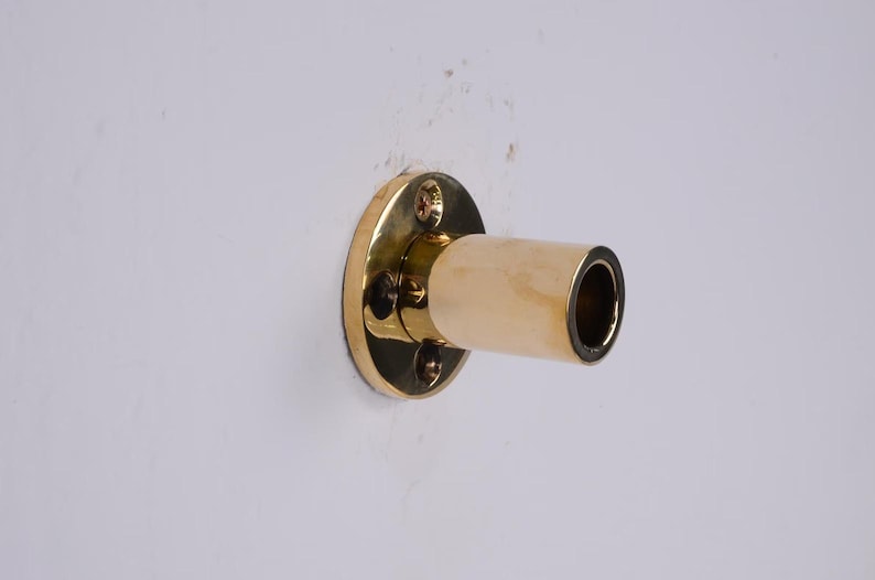 May include: A brass-colored wall-mounted curtain rod holder. The holder has a circular base with four screws and a cylindrical extension. The extension is a light beige color with a dark interior.