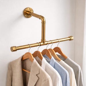 Puede incluir: Un perchero de ropa de estilo industrial dorado, montado en una pared blanca. El perchero está hecho de tuberías y accesorios, con una barra horizontal que sostiene perchas de madera con varias prendas, incluyendo una blazer beige y camisas.