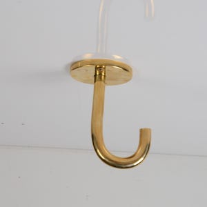 May include: A brass-colored ceiling hook with a curved design. The hook is attached to a circular base and a clear, cylindrical stem. The hook is designed to be mounted on a ceiling.