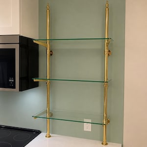 May include: A wall-mounted shelving unit featuring two vertical polished brass poles with decorative finials and three horizontal clear glass shelves supported by brass brackets. The structure is secured to the wall with circular brass mounts.