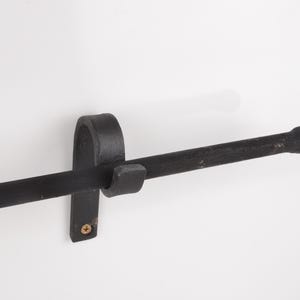 May include: A black, wrought iron towel bar mounted on a white wall. The bar is horizontal with a rounded end and is held in place by a curved bracket and a small, square piece. A single screw secures the bracket.