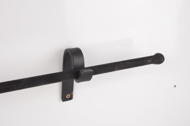 May include: A black, wrought iron towel bar mounted on a white wall. The bar is horizontal with a rounded end and is held in place by a curved bracket and a small, square piece. A single screw secures the bracket.