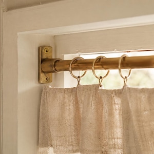 Könnte beinhalten: Eine Messing-Gardinenstange, die mit einer rechteckigen Halterung und zwei Schrauben an einem weißen Fensterrahmen befestigt ist. Die Stange verfügt über vier runde Messingringe, die einen gerafften, cremeweißen, strukturierten Vorhang halten.