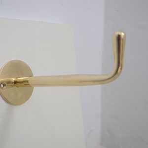 Może przedstawiać: Mosiężny hak ścienny z zaokrąglonym, zakrzywionym ramieniem. Hak jest zamontowany na okrągłej podstawie za pomocą jednego wkręta.