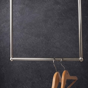 May include: A chrome-finished clothing rack with two wooden hangers suspended from the bar. The rack has a rectangular frame and is set against a dark, textured background. The hangers are made of light-colored wood.