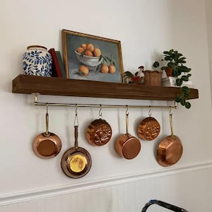 Może przedstawiać: Drewniana półka kuchenna z wiszącymi miedzianymi patelniami. Półka zawiera obraz pomarańczy, niebiesko-biały wazon, książki, figurkę koguta i roślinę doniczkową. Patelnie są w różnych rozmiarach i kształtach.