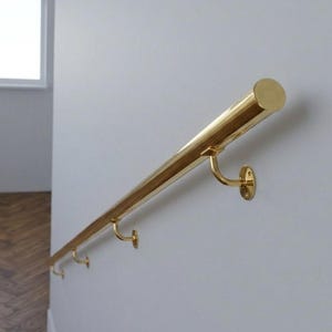 Puede incluir: Un pasamanos de latón pulido montado en una pared blanca. El pasamanos es cilíndrico con soportes curvos. El latón tiene una superficie brillante y reflectante. El pasamanos mide unos 100 cm de largo.